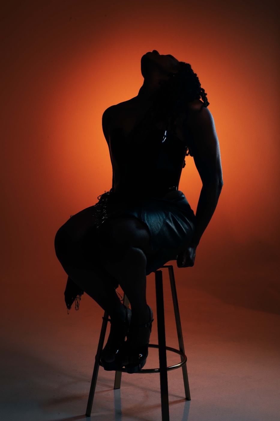 Domina Divinia in silhouette, seated on a stool, rim-lit against a dark background.